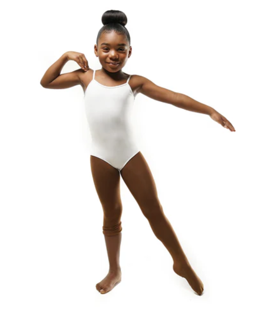 A happy young Black girl in a bun, wearing a white leotard and medium brown transition tights the color “brazen brown”. One leg is up to the knee and the other over her foot.